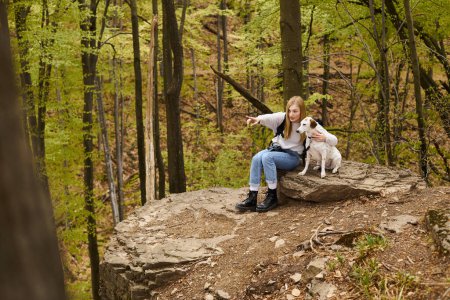 Aventurera rubia devota mostrando a su curioso perro una dirección sentada en la roca en el bosque