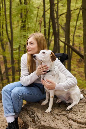 Téléchargez les photos : Femme blonde souriante étreignant son chien, tout en s'arrêtant en voyage en forêt, les deux regardant ailleurs - en image libre de droit