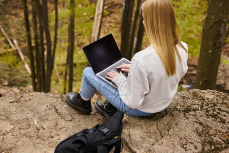 Back view of blonde female hiker sitting on boulder in forest with laptop on her knees with backpack