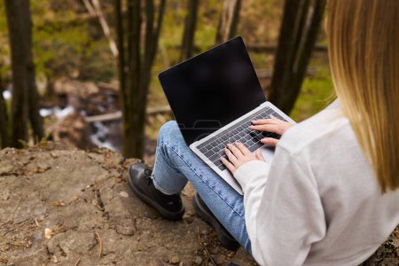 Téléchargez les photos : Vue arrière d'une femme blonde assise sur un grand rocher dans la forêt, utilisant un ordinateur portable pour un travail à distance - en image libre de droit