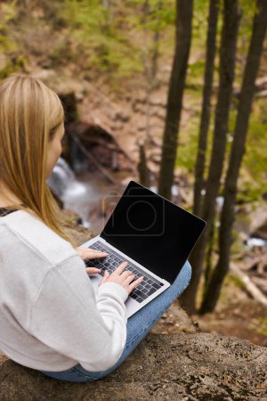Blondine mit Rucksacktour und Ruhepause auf Felsbrocken im Wald, arbeitet am Laptop