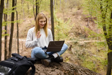 Mujer rubia joven con mochila en el bosque, sentada en la roca con el ordenador portátil y haciendo videollamadas