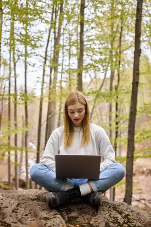 Blonde, entspannte Frau im Wald, sitzt auf Felsbrocken mit Laptop in Lotusposition