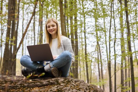 Smiling bright woman on halt in woods, sitting on boulder with laptop in lotus position