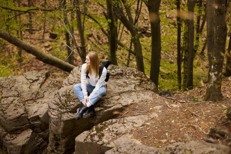 Young woman hiker with backpack sitting at halt on rocky cliff in forest looking away
