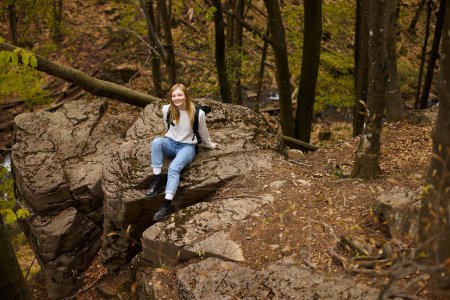 Smiling woman hiker with backpack sitting at halt on rocky cliff in forest looking at camera