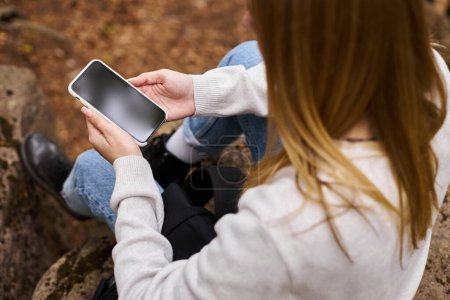 Téléchargez les photos : Image recadrée de la femme tenant le téléphone dans les mains assis dans les paysages forestiers et textos - en image libre de droit