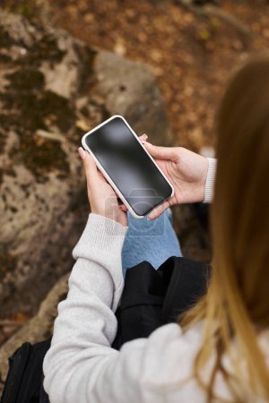 Zugeschnittenes Bild einer Frau, die ihr Handy in der Hand hält, in einer Waldlandschaft sitzt und auf den Bildschirm schaut