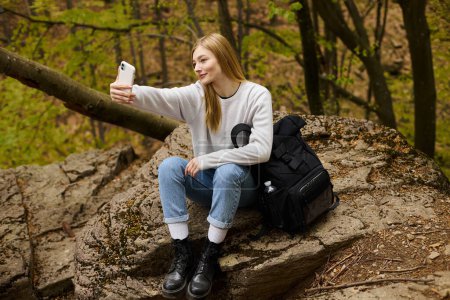 blonde young woman taking selfie sitting on rock at halt in the forest while hiking