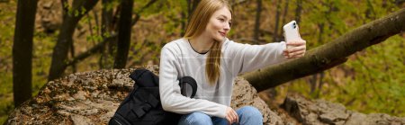 mujer joven rubia tomando selfie sentado en la roca en alto en el bosque mientras caminaba, pancarta