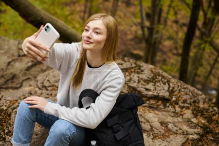Foto de Sonriendo tranquila joven rubia tomando selfie mientras se relaja en el bosque mientras camina - Imagen libre de derechos
