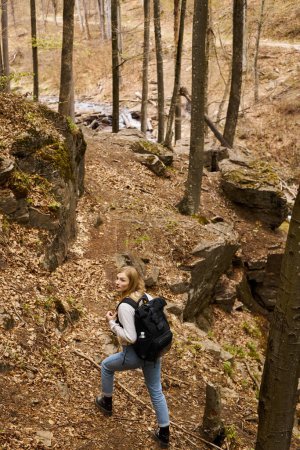 Back view of cheerful blonde female hiker wearing backpack crossing the forest with stream