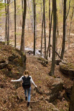 Back view of young blonde female traveler wearing backpack running to forest stream