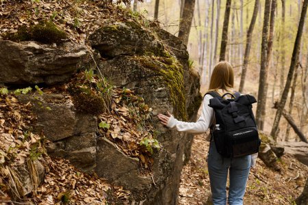 Vista trasera de la mujer rubia viajera con mochila en viaje de senderismo y caminando cerca de acantilado rocoso