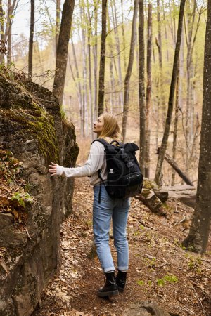 Vista trasera de la curiosa mujer rubia con mochila en viaje de senderismo y caminando cerca de acantilado rocoso
