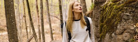 Adventurous blonde woman wearing backpack on hiking trip and walking near rocky cliff, banner