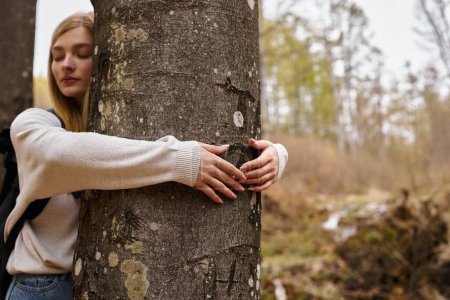 Entspannte junge blonde Wanderin mit Reiserucksack, umarmtem Baum und friedlichem Ausflug