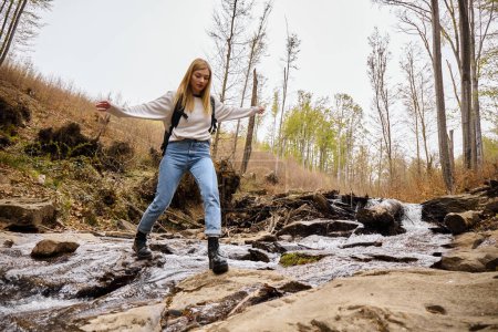 Joyful blonde female hiker wearing sweater and jeans crossing the forest creek jumping over water