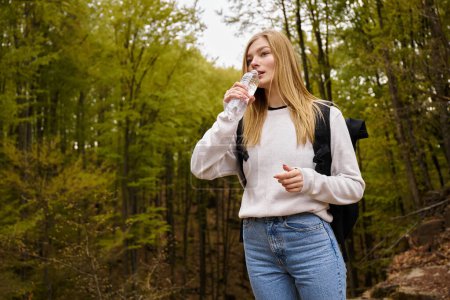 Woman traveler wearing sweater and jeans crossing the forest creek walking in woods drinking water