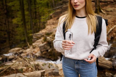 Mujer viajera vistiendo suéter y jeans cruzando el bosque sosteniendo botella de agua