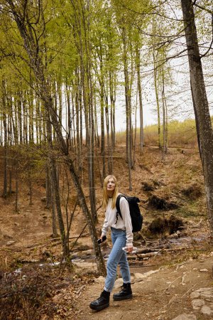 Young woman traveler wearing sweater and jeans crossing the forest holding bottle of water