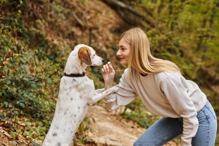 Cheerful woman training her pet dog holding leash at hiking rest with mountain and forest view