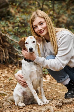 Cheerful woman loving and hugging gently her dog and looking at camera while walking in woods