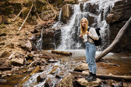 Active blonde female hiker standing by the forest creek walking on rocks near waterfall