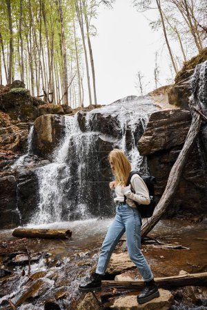 Active blonde female hiker crossing the forest creek walking on rocks near waterfall