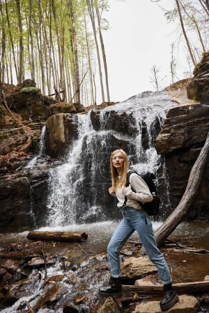 Young female hiker crossing the forest creek, having backpack trip near waterfall
