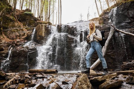Young blonde female hiker crossing the forest creek walking on rocks near waterfall