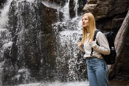 Portrait of young blonde female traveler hiking in beautiful forest and standing near waterfall