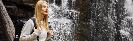 Portrait of young blonde female traveler hiking in forest and standing near waterfall, banner