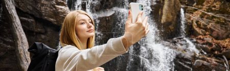 Foto de Mujer joven rubia tomando selfie cerca de la cascada de montaña en el bosque mientras caminaba, pancarta - Imagen libre de derechos