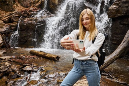Foto de Sonriente joven rubia tomando selfie cerca de la cascada de montaña en el bosque mientras caminaba - Imagen libre de derechos