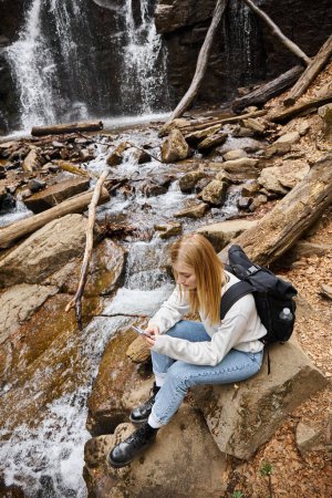 blondynka kobieta podróżnik za pomocą telefonu siedzi w pobliżu górskiego wodospadu w lesie podczas wędrówki