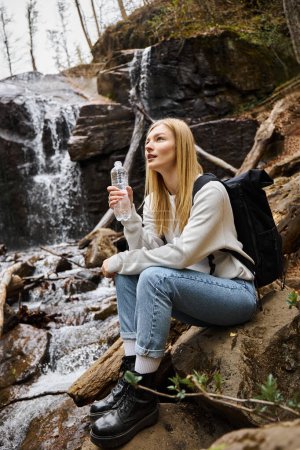 Młoda kobieta z plecakiem siedząc i trzymając butelkę wody i patrząc na las w pobliżu wodospadu