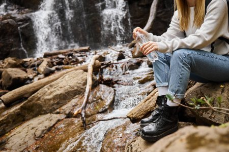 Foto de Imagen recortada de mujer en jeans sentada con botella de agua sobre rocas en el bosque cerca de la cascada - Imagen libre de derechos