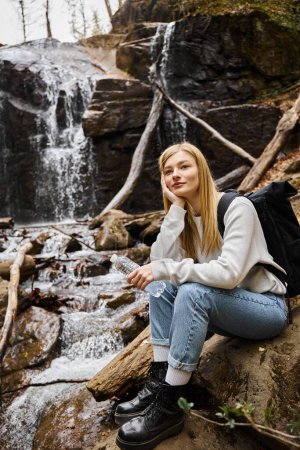 Foto de Mujer sonriente sentada con botella de agua y mirando hacia otro lado en el bosque cerca de la cascada - Imagen libre de derechos