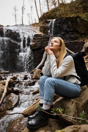 Foto de Mujer soñadora sentada con mochila y mirando al cielo en el bosque cerca de la cascada - Imagen libre de derechos