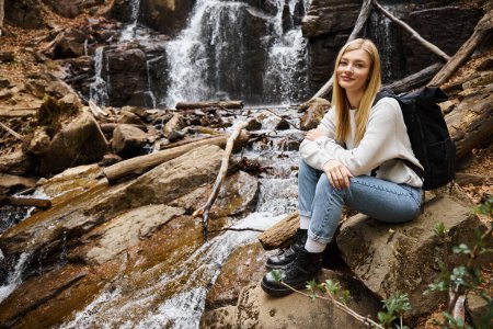 Mujer aventurera excursionista sentado con mochila y mirando a la cámara en el bosque cerca de la cascada