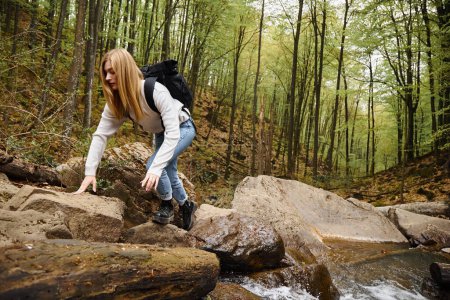 Woman with backpack hiking and crawling on mountain river rock in autumn forest. Solo female tourist