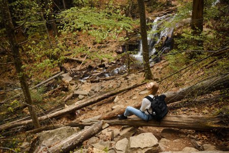 Widok z tyłu młoda blondynka turystka siedzi w pobliżu wodospadu i odpoczynku podczas trekkingu, przygoda