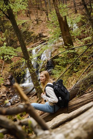 Foto de Vista trasera de la joven senderista rubia sentada cerca de la cascada y descansando, mirando hacia otro lado - Imagen libre de derechos