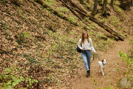 Young woman walking her pet dog holding leash at hiking rest with mountain and forest view