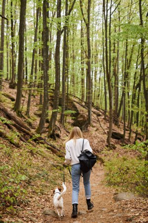 Back view of blonde young active woman walking her dog companion at forest backpacking trip