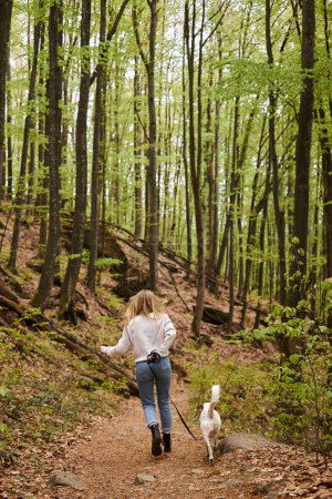 Back view of active blonde woman running with her cute white dog while hiking in forest