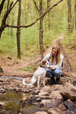 Smiling woman hugging gently her dog companion and looking at pet while walking in forest