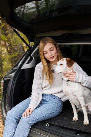 Blonde junge Frau in Pullover und Jeans umarmt ihren Hund, der beim Wandern im Wald im Auto sitzt