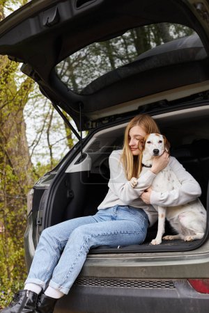 Smiling happy woman hugging her dog sitting in back of car in forest at hiking trip halt